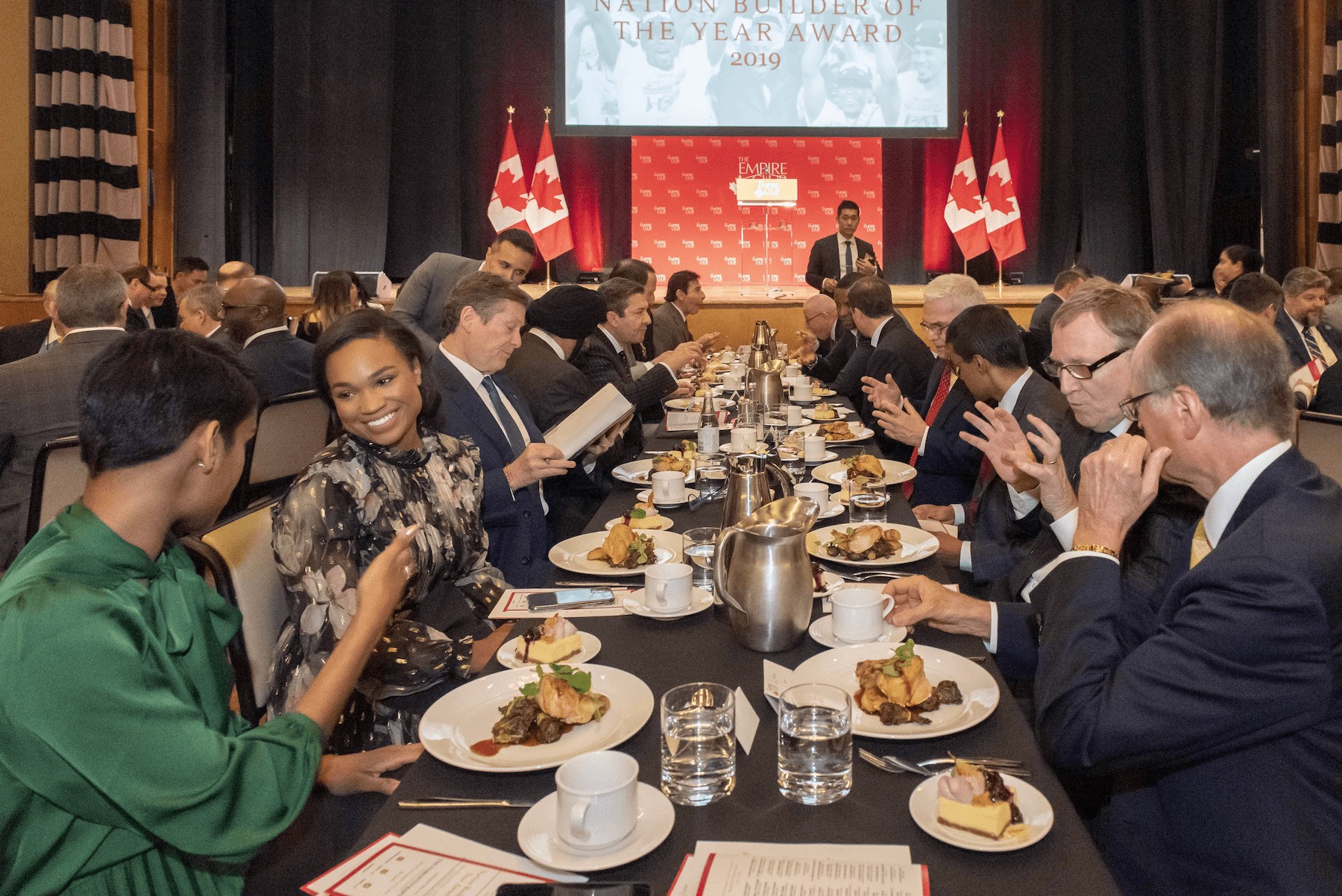 alt= Smiling professional woman sitting at at an event table with food and other Nation Builder 2019 attendees.
