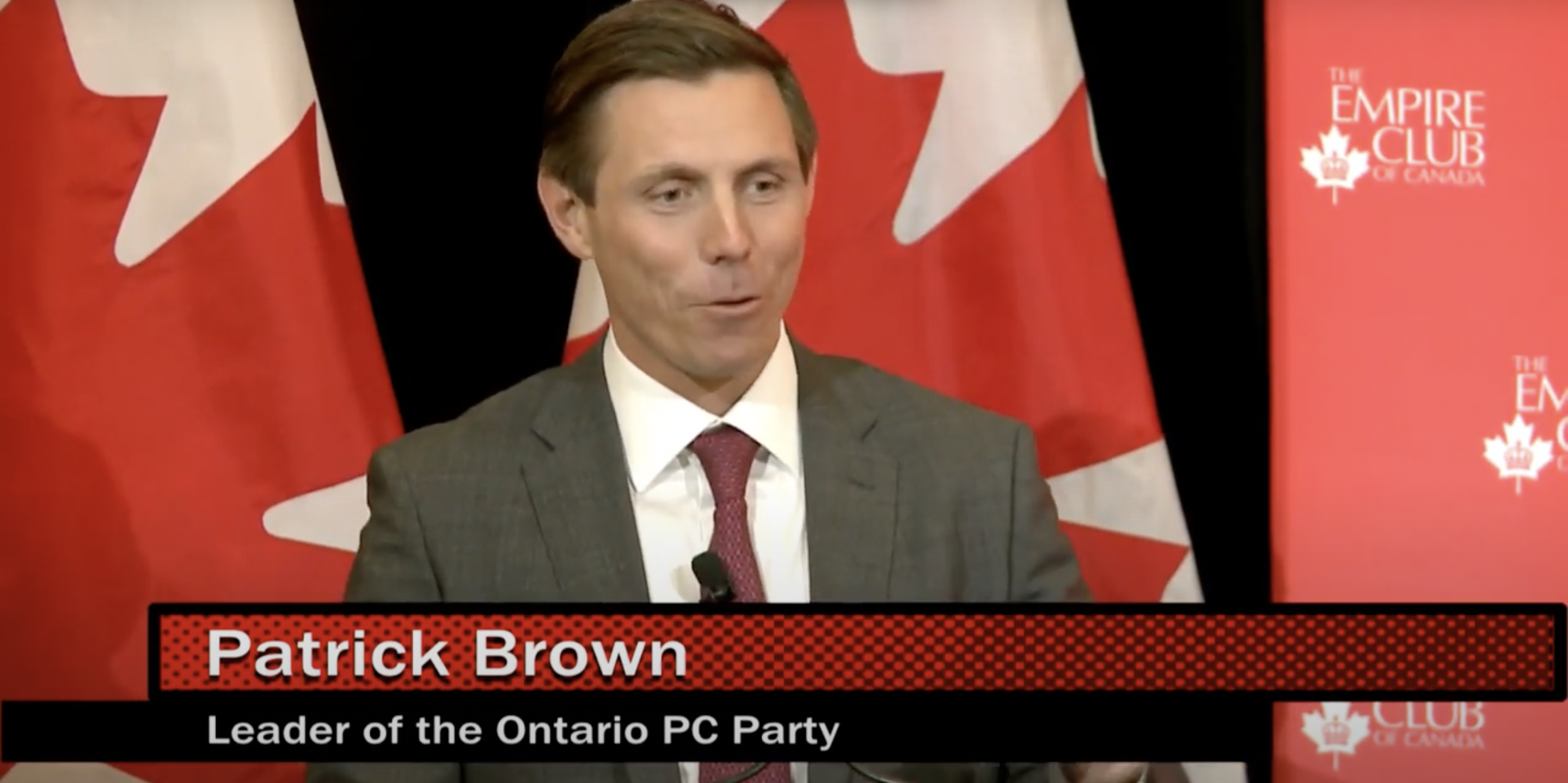 alt= A man, Patrick Brown, standing and speaking in front of Canadian flags
