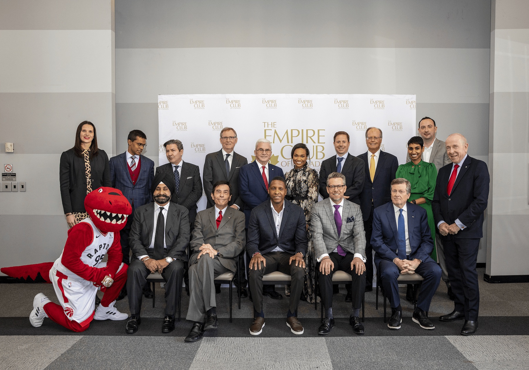 alt= Empire Club of Canada Directors and members smiling with the Toronto Raptors red raptor mascot in front of an event wall.