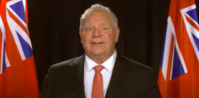 alt= Man, 26th Premier of Ontario, wearing black suit standing between flags.