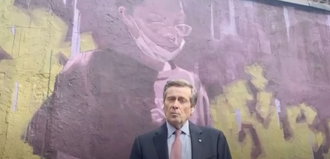 alt= Mayor of Toronto, John Tory, wearing a blue suit and standing in front of outdoor street art.