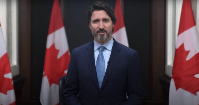 alt= Man, 23rd Prime Minister of Canada, wearing a blue suit and tie standing with several Canadian flags.