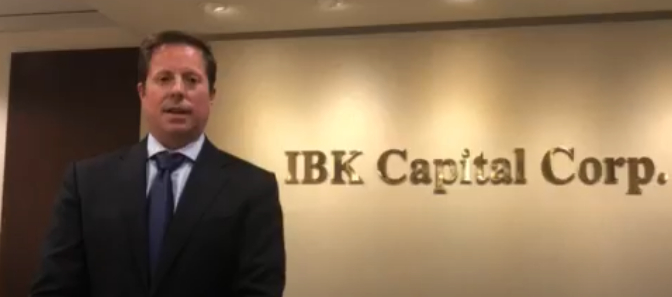 alt= Man wearing a black suit standing in front of a wall with "IBK Capital Corp." on it.