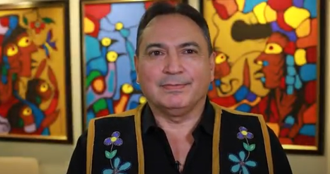 alt= Man wearing a black shirt with floral designs standing in front of color art pieces hung on the wall.