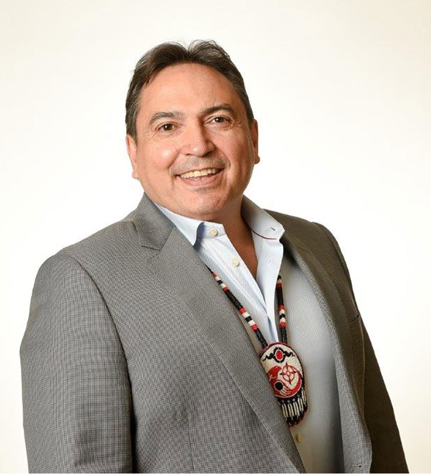 alt= Former National Chief of the Assembly of First Nations, Perry Bellegarde, smiling and wearing a grey suit with a First Nations neck piece.