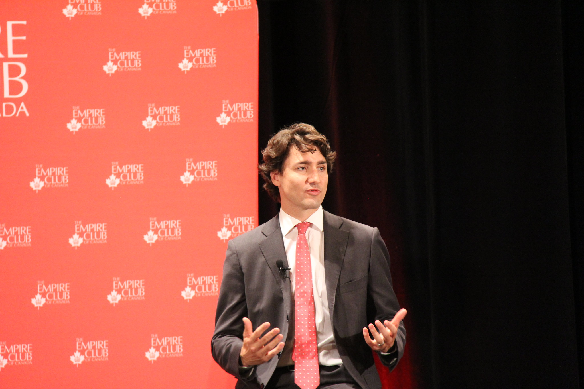 Prime Minister Justin Trudeau speaking with open hands. He is wearing a dark suit jacket, white shirt and red tie and is standing in front of a red background