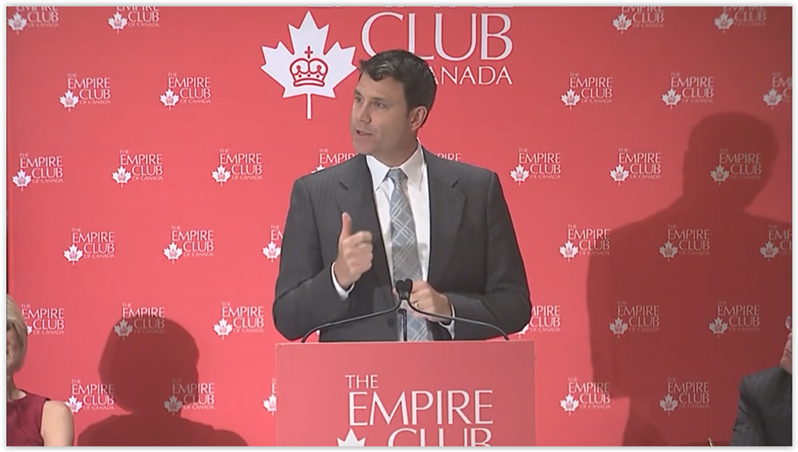 a man in a grey suit delivering a keynote speech at the Empire Club of Canada podium