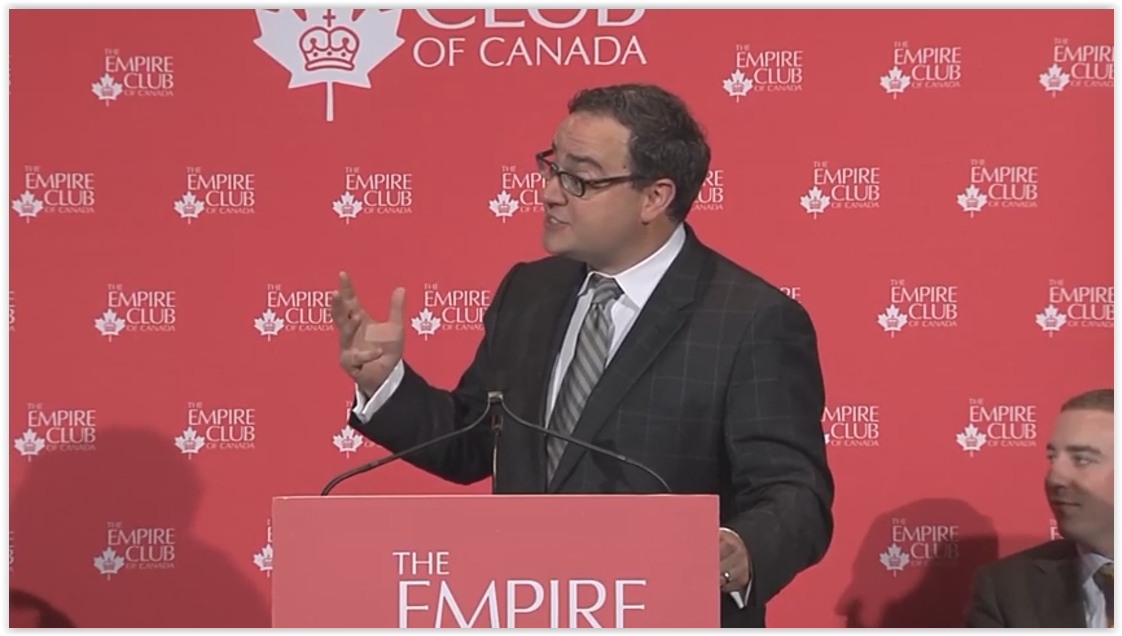Man in a grey suit jacket gesticulating with his hand as he delivers a speech from the podium at the Empire Club of Canada