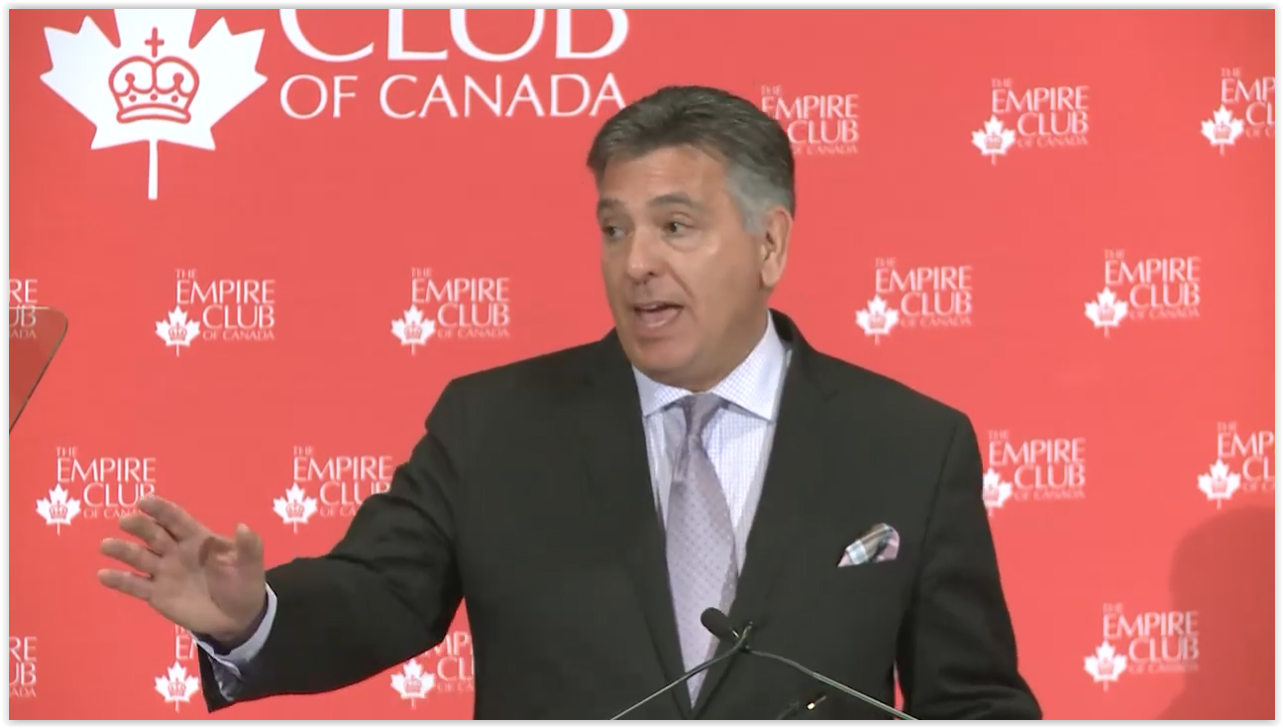Man in a suit with a white shirt and lilac tie speaking at the podium in front of a red background with white Empire Club logo